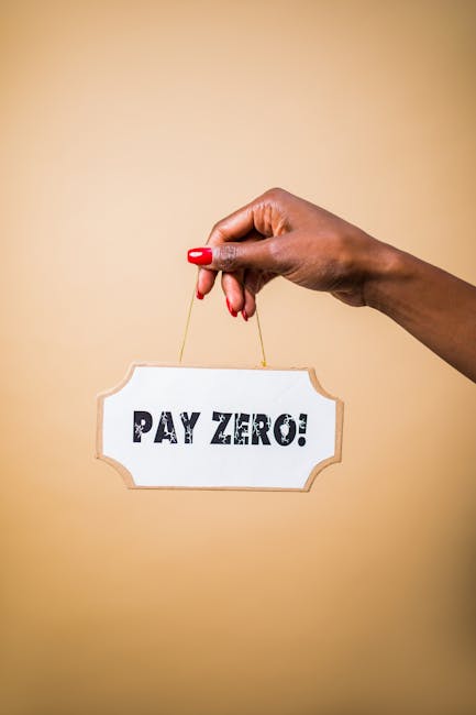 A close-up of a person’s hand with red nail polish holding a small, rectangular sign by a thin string. The sign reads 'PAY ZERO!' in bold black capital letters, with a slightly decorative border. Th