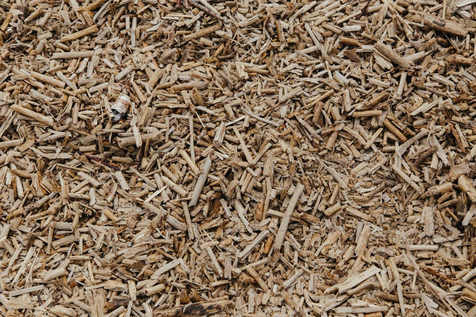 A close-up view of a garden bed filled with finely chopped wood mulch, showcasing a mixture of small, irregularly shaped pieces with rough textures and natural wood tones. The mulch covers the soil su