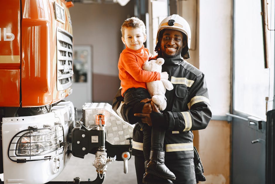 A firefighter in a black uniform and helmet is holding a young child with curly hair, dressed in an orange sweater and black pants, inside a fire station. The child is smiling and holding a white tedd